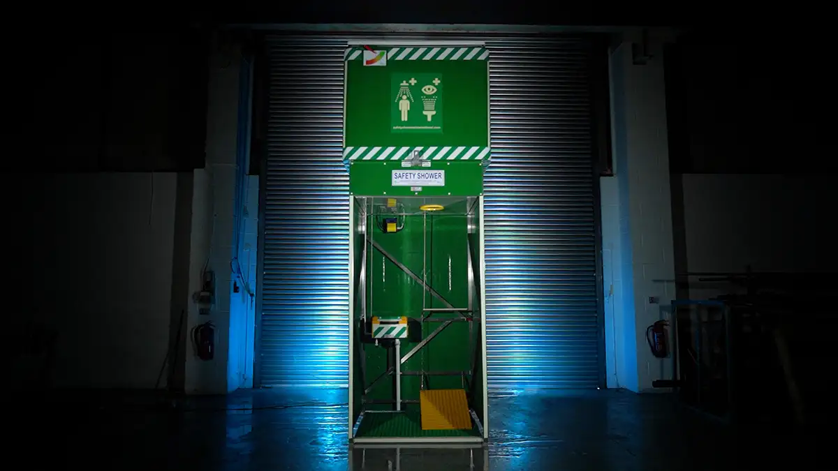A green Aqua safety shower against a stylised industrial backdrop. The emergency shower features a 1500L header tank on a stainless steel frame, with an internal eye wash station and foot platform activation.